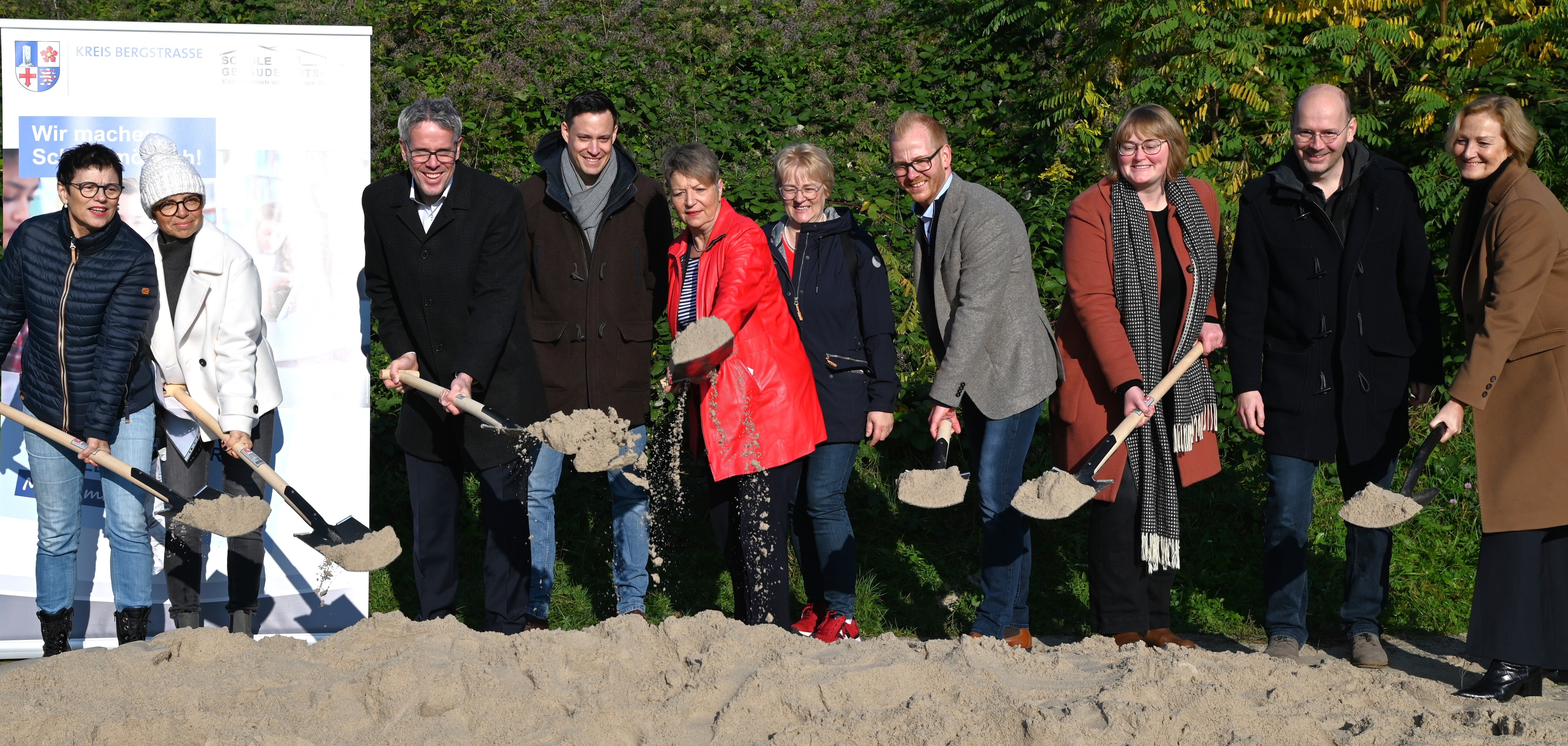 Die Ortsvorsteherin von Bensheim-West, Ingrid Schich-Kiefer, die Schulleiterin Helga Krumbein, Landrat Christian Engelhardt, der kaufmännische Leiter des Eigenbetriebs Schule und Gebäudewirtschaft, Simon Menden, Bürgermeisterin Christine Klein, die Vertreterin des Kreiselternbeirats, Daniela Jahn, der technischer Leiter des Eigenbetriebs Schule und Gebäudewirtschaft, Johannes Kühn, Anne Kettenburg von Werk.um Architekten, Christian Keil und Christine Grimm von Mobispace GmbH stehen auf dem Gelände des Neubaus vor einem Sandhügel und führen mit Schaufeln den symbolischen Spatenstich aus.