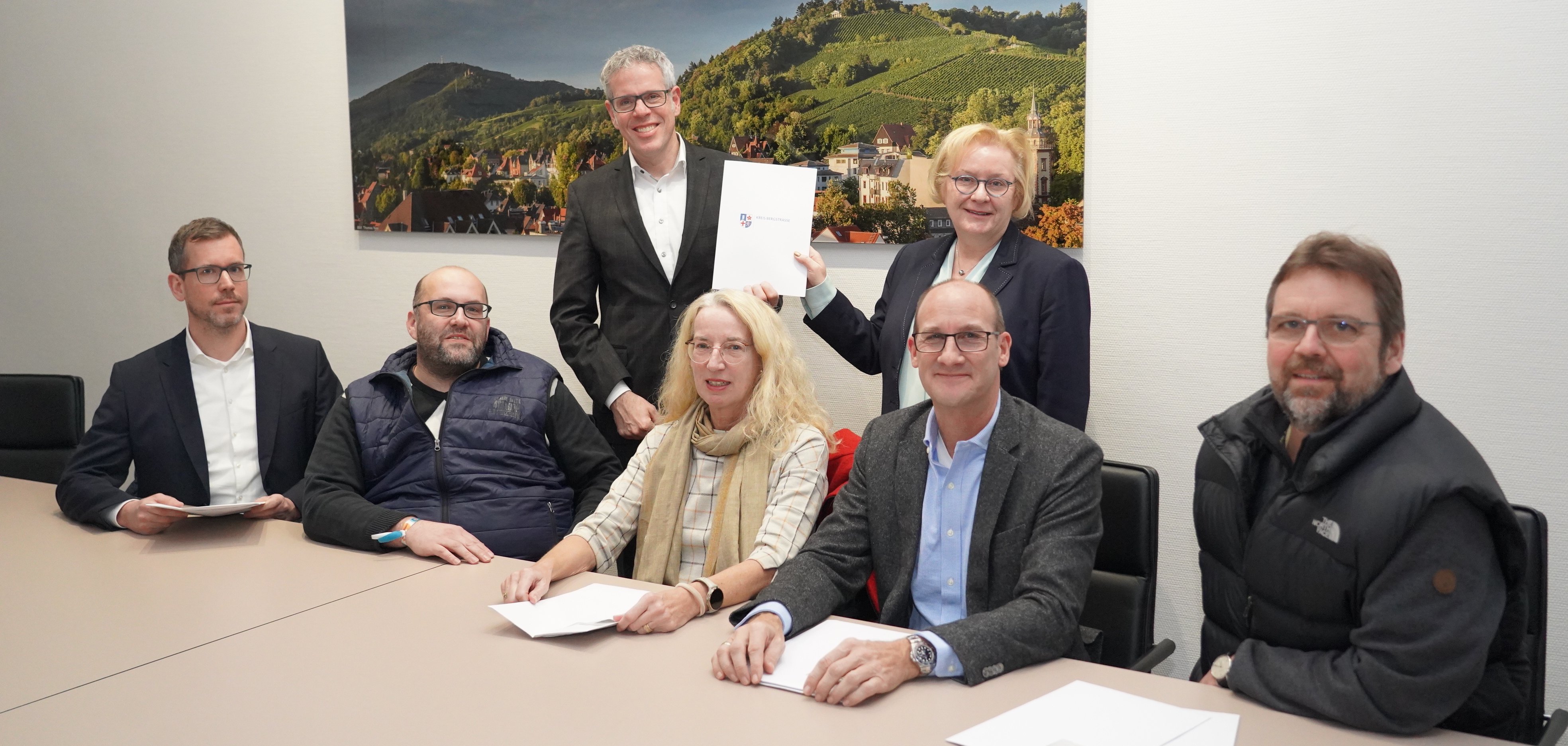 Landrat Christian Engelhardt und die Erste Stadträtin Bensheims, Nicole Rauber-Jung halten gemeinsam den Förderbescheid. Für ihnen sitzen an einem Tisch Stellvertretend für die Ortsteile Dr. Stefan Hebenstreit (Ortsvorsteher Gronau), Michael Gortz (Ortsvorsteher Schönberg), Sabine Hinterkeuser-Feye (Ortsvorsteherin Hochstädten), Konrad Klapfenberger (Ortsvorsteher Schwanheim) sowie Dr. Stefan Stötzel (Ortsvorsteher Fehlheim).