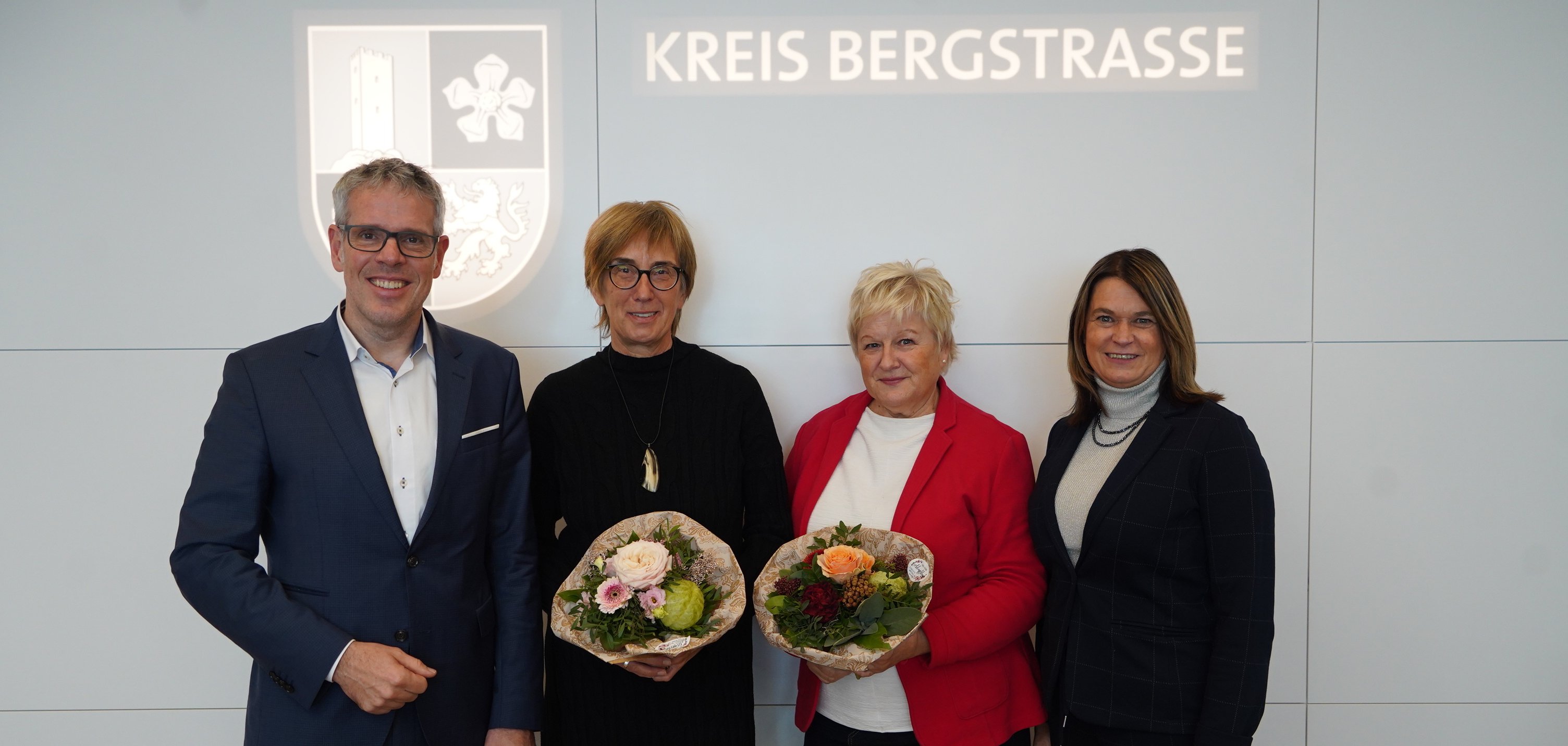 Landrat Christian Engelhardt und die Erste Kreisbeigeordnete Angelika Beckenbach stehen mit Claudia Blume und Karin Ende vor einer Wand mit dem Logo des Kreises Bergstraße darauf. Die beiden Kolleginnen halten Blumensträuße in den Händen.