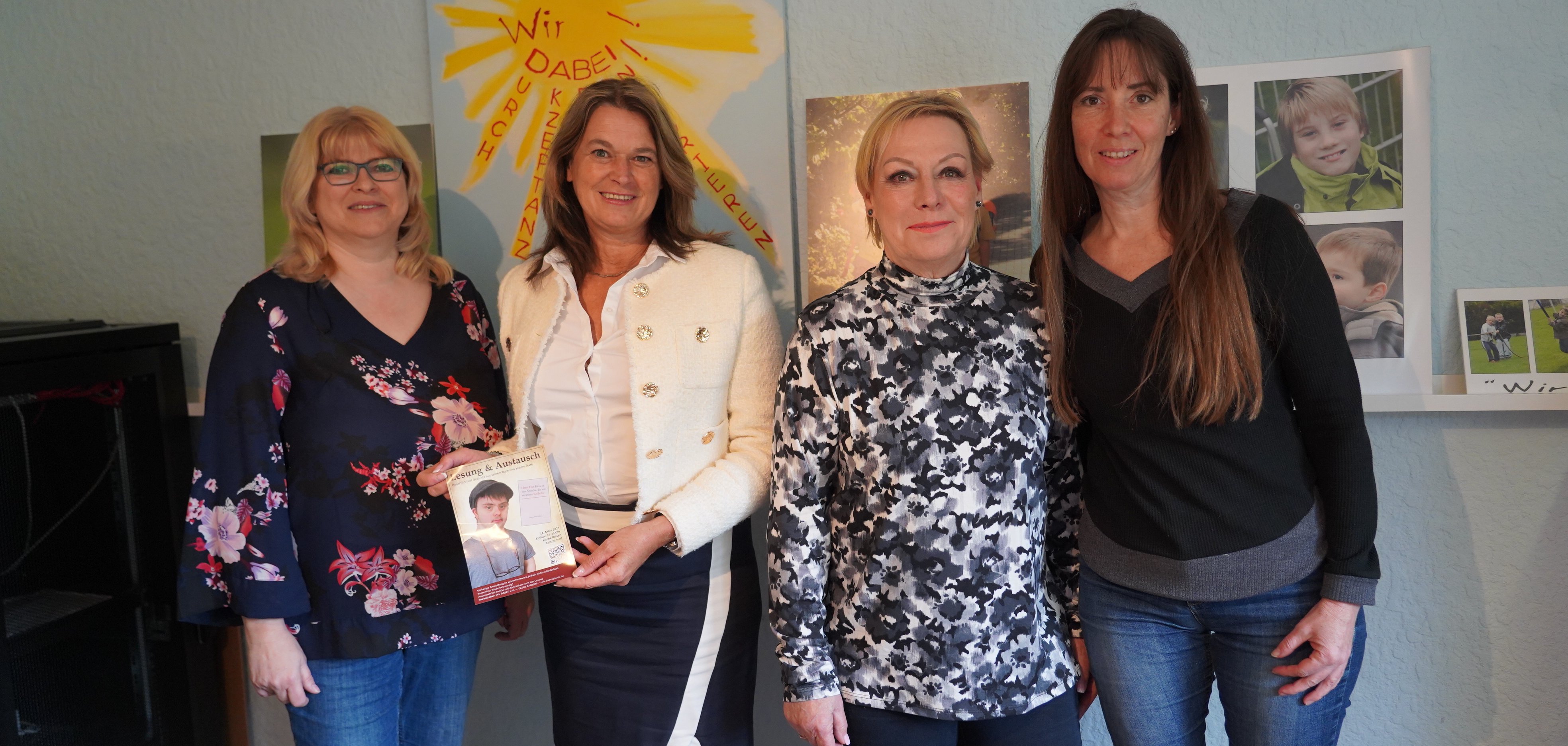 Die Erste Kreisbeigeordnete Angelika Beckenbach, die Leiterin der EUTB Bergstraße-Odenwald, Petra Thaidigsmann , die Beraterin Petra Doering der allgemeinen Beratungsstelle und Heike Klemm von der Geschäftsstelle des Vereins Wir DABEI! e.V. stehen vor einer Wand, auf der eine Sonne gemalt ist. Angelika Beckenbach hält einen Flyer zur Lesung in den Händen.