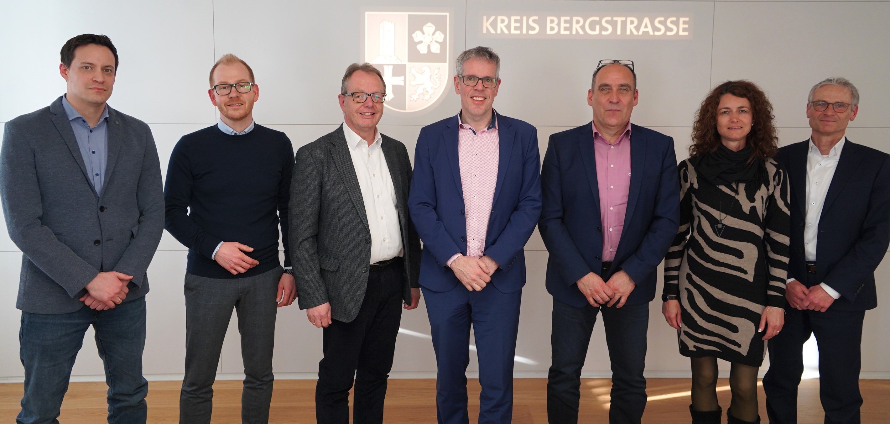 Die Unterzeichnenden der Kooperationsvereinbarung stehen im Landratsamt in einem Besprechungssaal vor einer Wand mit dem Logo des Kreises Bergstraße nebeneinander. Vor ihnen liegt auf einem Tisch der Vertrag.