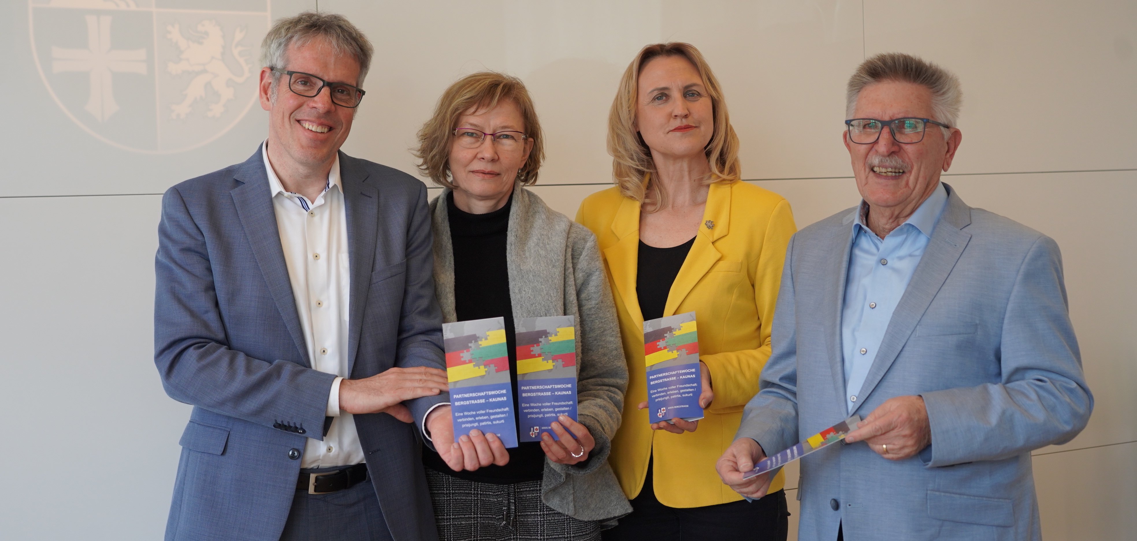 Landrat Christian Engelhardt steht mit Ewa Redemann von der Abteilung Bürgerservice, Vereine und Kultur des Kreises Bergstraße, der Leiterin des Litauischen Gymnasiums Lampertheim-Hüttenfeld, Rasa Weiß sowie dem Kreisbeigeordneten und Leiter des Arbeitskreises „Forum Kaunas“, Heinz Klee in einem Besprechungssaal vor einer Wand mit dem Logo des Kreises Bergstraße darauf. Sie alle halten das Programm der diesjährigen Partnerschaftswoche Bergstraße-Kaunas in den Händen.