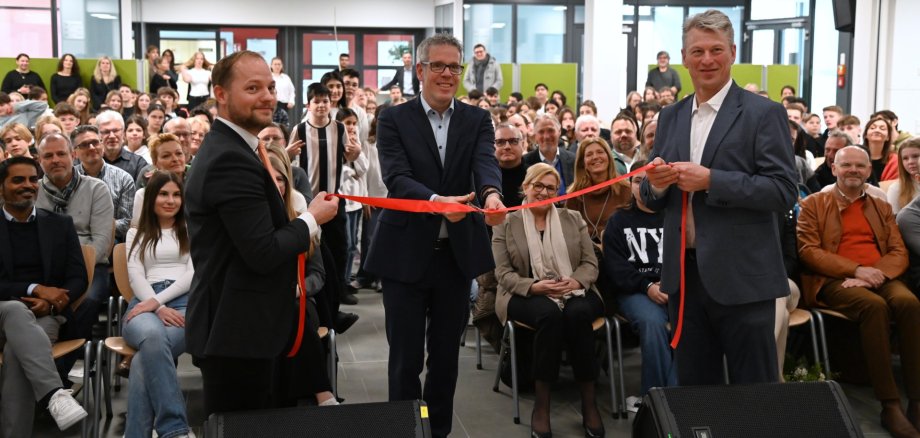 Schulleiter Arndt Neumann und sein Stellvertreter Thomas Rech halten vor der Schulgemeinde das rote Band für den offiziellen Schnitt durch Landrat Christian Engelhardt.