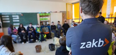 Gabi Köhler vom ZAKB steht mit dem Rücken zur Kamera vor einem Kreis aus Schülerinnen und Schülern.