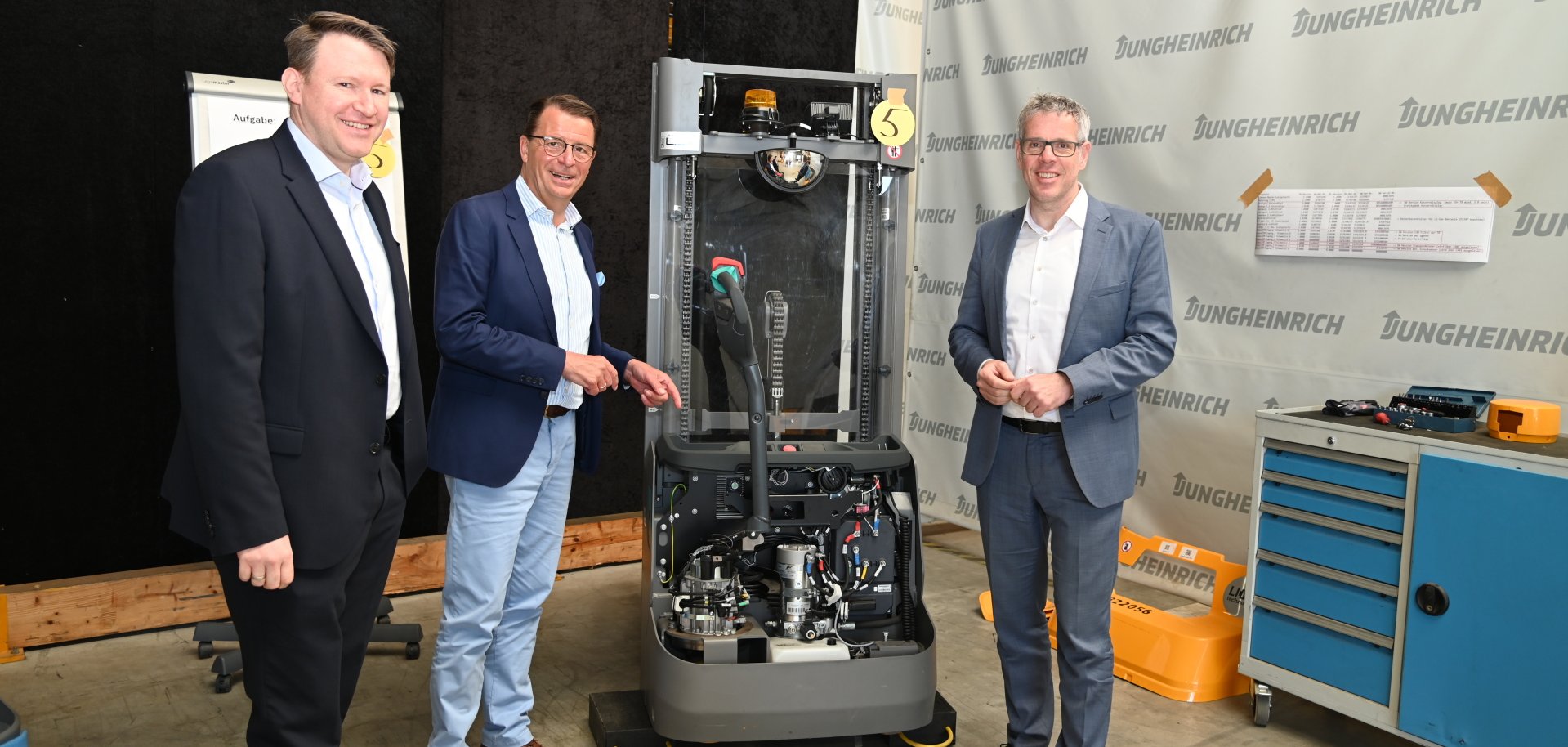 Dr. Matthias Zürker, Geschäftsführer der Wirtschaftsförderung Bergstraße, der Leiter des Vertriebszentrum Südwest von Jungheinrich, Uwe Seitz, und Landrat Christian Engelhardt stehen in der Werkstatt des Unternehmens und zeigen auf einen Gabelstapler.