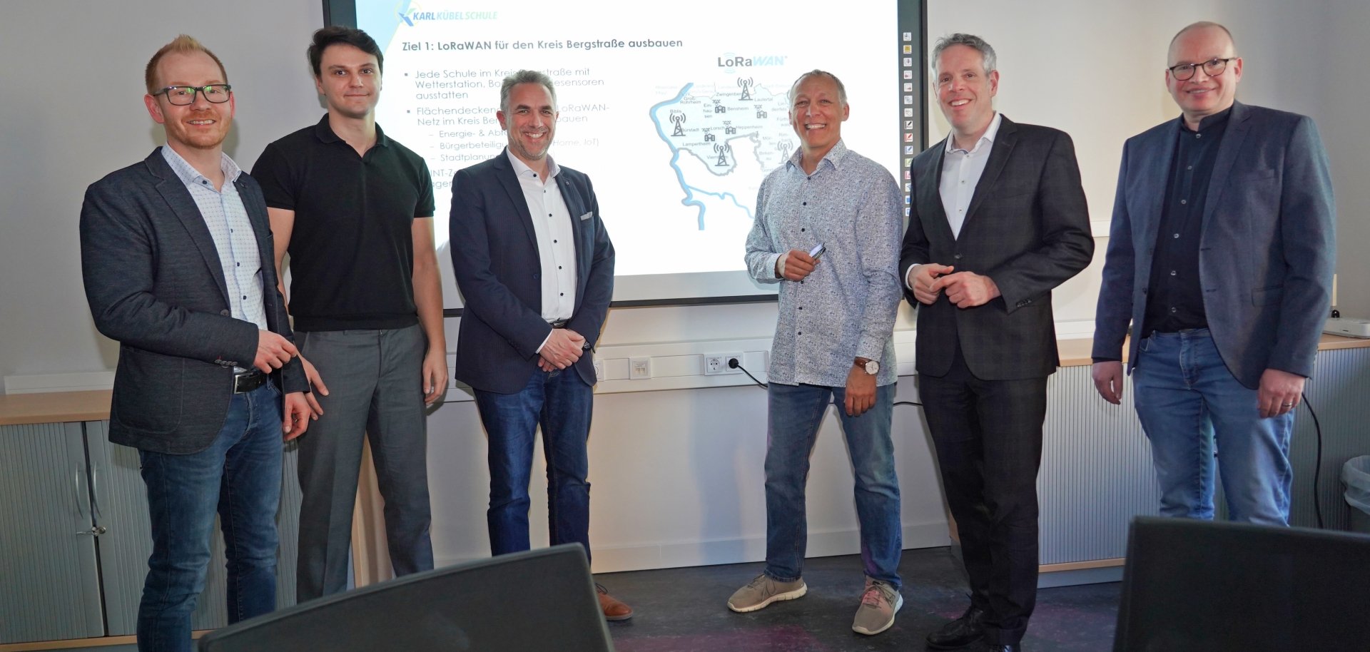 Landrat Christian Engelhardt,  Johannes Kühn, Abteilungsleiter des Eigenbetriebs Schule und Gebäudewirtschaft des Kreises Bergstraße, Navin Dass, Schulleiter Michael Steffan, Rainer Wieland und Thorsten Waizmann von der Karl Kübel Schule stehen nebeneinander vor einem Smartboard, auf dem die Titel der Klimdatenprojekte abgebildet sind.