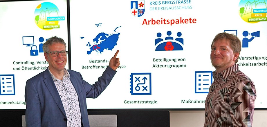 Landrat Christian Engelhardt (links) und Klimaanpassungsmanager Alexander Uhl (rechts) zeigen auf einen Monitor hinter ihnen, auf denen Aspekte des Klimaanpassungskonzepts abgebildet sind.