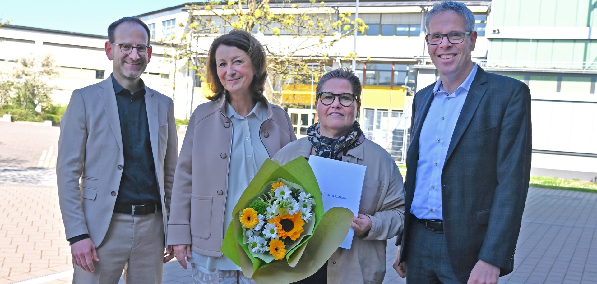 Landrat Christian Engelhardt steht gemeinsam mit Sabine Karb, der Schulleiterin Silke Weimar-Ekdur und dem stellvertretenden Schulleiter Jérôme Dath nebeneinander vor dem Schulgebäude. Frau Karb hält einen Blumenstrauß in den Händen.