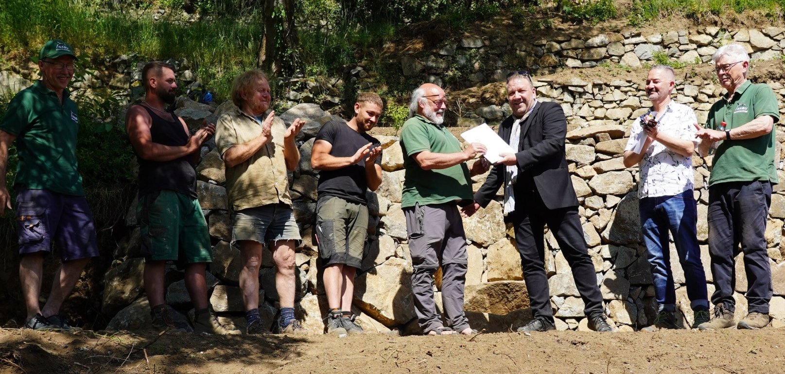 Kreisbeigeordneter Matthias Schimpf (3. v. r.) überreicht den Förderbescheid zur Sanierung der Trockenmauer, mit dabei (v.l.n.r.): Friedrich Jung (Beisitzer des NABU Meerbachtal), Thorsten Navratil, Thomas Müller, Lutz Müller, Jürgen Schneider (Vorsitzender des NABU Meerbachtal), Michael Weidner (Untere Naturschutzbehörde) und Werner Hombeuel (2. Vorsitzender des NABU Meerbachtal), während sie auf einem Bauabschnitt des Geländes stehen.