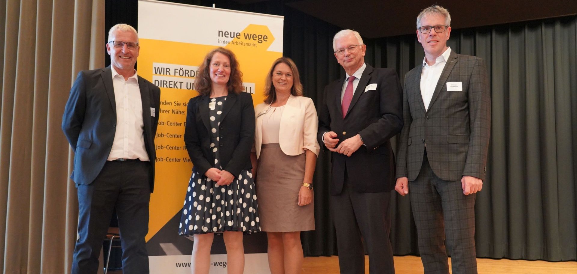 Landrat Christian Engelhardt, der Hessische Ministerpräsident a.D., Prof. Dr. Roland Koch, die Erste Kreisbeigeordnete Angelika Beckenbach, die Leiterin des Eigenbetriebes Neue Wege -Kommunales Jobcenter- Kreis Bergstraße, Dr. Melanie Marysko, sowie ihr Stellvertreter Peter Schmiedel stehen bei der Jubiläumsfeier in Mörlenbach nebeneinander auf der Bühne vor einem Neue Wege Roll-Up.