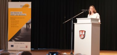 Begrüßung Erste Kreisbeigeordnete Die Erste Kreisbeigeordnete und für das Thema zuständige Dezernentin Angelika Beckenbach steht auf der Bühne an einem Podium.