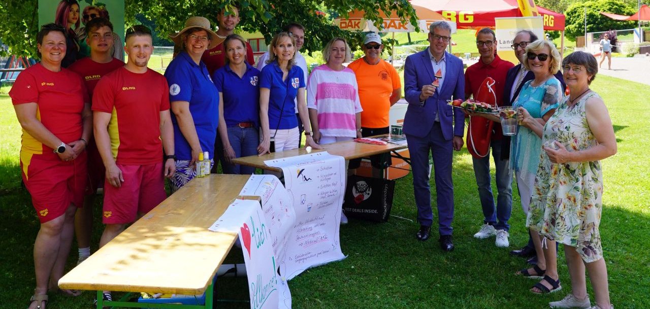 Landrat Christian Engelhardt (5. v.r.) posiert zusammen mit (v.l.n.r.) Vertreterinnen und Vertretern des DLRG Ortsverbandes Wald-Michelbach, Ute Fuchs vom Gesundheitsamt des Kreises, Reinhild Zolg und Nadja Niestroj vom Präventionsteam des Kreises, Robert Gahler und Verena Burgmer vom DAK-Gesundheit Servicezentrums Bensheim, Schwimmmeister Wolf-Dieter Schmidt sowie Apotheker Rolf Schaffnith, dem Bürgermeister von Wald-Michelbach, Dr. Sascha Weber und Annette Folz und Claudia Jöst vom Bezirkslandfrauenverein Heppenheim auf einer grünen Wiese im Waldschwimmbad Wald-Michelbach für den Sonnenschutz Aktionstag