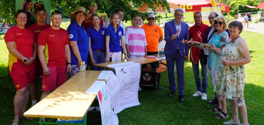 Landrat Christian Engelhardt (5. v.r.) posiert zusammen mit (v.l.n.r.) Vertreterinnen und Vertretern des DLRG Ortsverbandes Wald-Michelbach, Ute Fuchs vom Gesundheitsamt des Kreises, Reinhild Zolg und Nadja Niestroj vom Präventionsteam des Kreises, Robert Gahler und Verena Burgmer vom DAK-Gesundheit Servicezentrums Bensheim, Schwimmmeister Wolf-Dieter Schmidt sowie Apotheker Rolf Schaffnith, dem Bürgermeister von Wald-Michelbach, Dr. Sascha Weber und Annette Folz und Claudia Jöst vom Bezirkslandfrauenverein Heppenheim auf einer grünen Wiese im Waldschwimmbad Wald-Michelbach für den Sonnenschutz Aktionstag