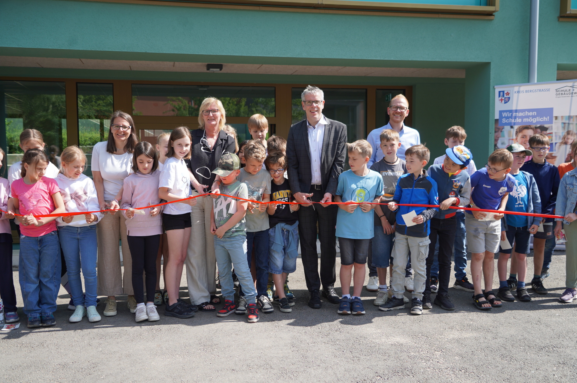 Landrat Christian Engelhardt (2. V. re.) schneidet gemeinsam mit der Ersten Stadträtin der Stadt Heppenheim Christine Bender, Schulleiterin Silke Jendrzejewski, dem technischen Betriebsleiter des Eigenbetriebes Schule und Gebäudewirtschaft Johannes Kühn (v.l.n.r.) sowie zahlreichen Schülerinnen und Schüler bei der Einweihung des neuen Verwaltungsgebäudes der Eichendorffschule Kirschhausen das rote Band durch.