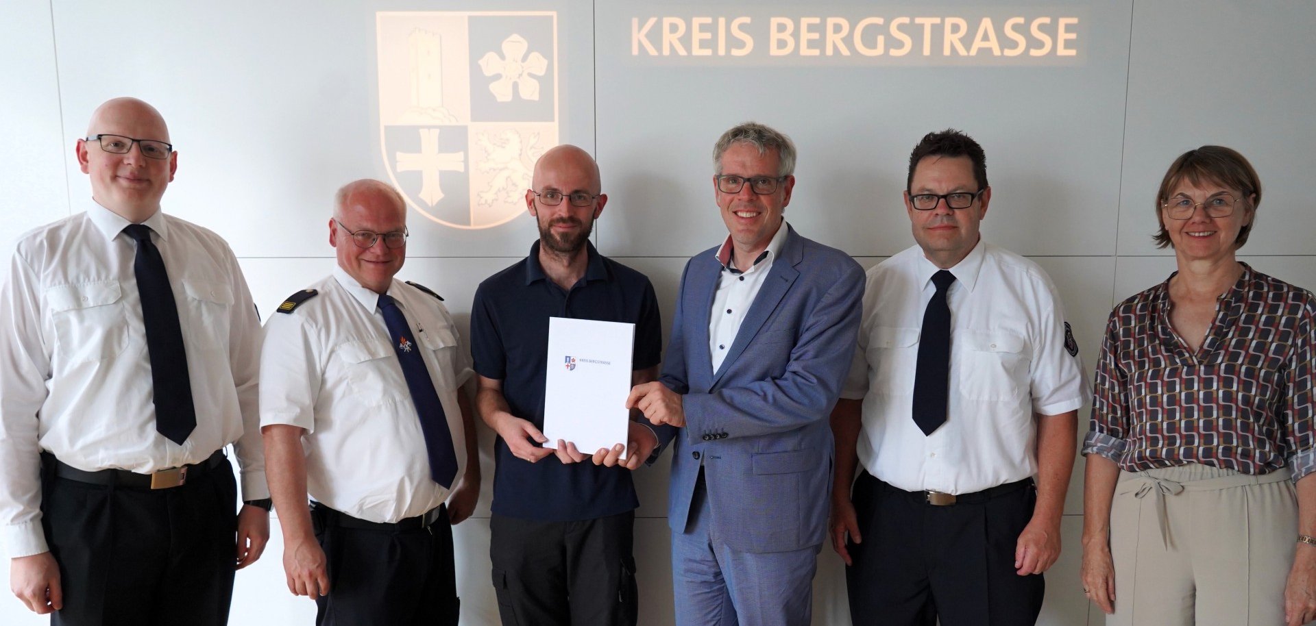 Landrat Christian Engelhardt steht mit Daniel Böhm, dem stellvertretenden Leiter der Abteilung Gefahrenabwehr des Kreises Markus Stracke, dem Leiter der Abteilung Gefahrenabwehr und Kreisbrandinspektor Stefan Lutter, dem Fachbereichsleiter Rettungsdienst in der Abteilung Gefahrenabwehr Jörg Oberkinkhaus und Barbara Hoffbauer, Leiterin der Abteilung Personalmanagement und Organisation in einem Besprechungssaal vor dem Logo des Kreises Bergstraße.