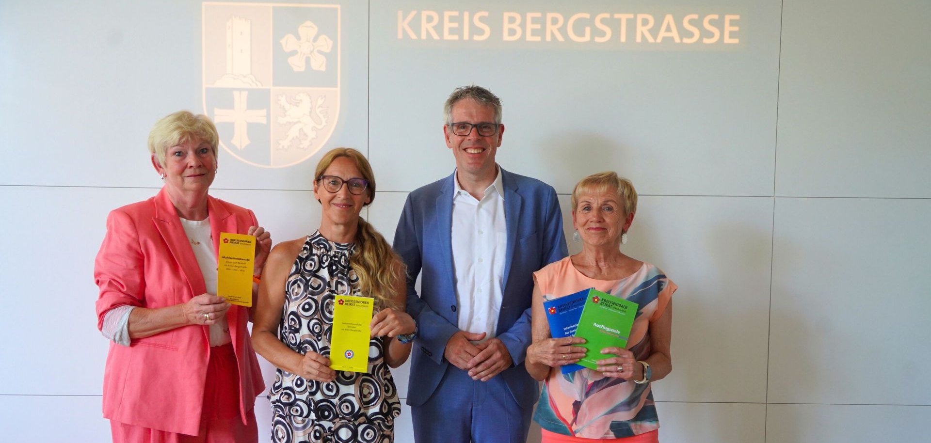 Landrat Christian Engelhardt steht mit der Vorsitzenden des Kreisseniorenbeirats, Susanne Hagen, der Vorsitzenden des Arbeitskreises „Seniorenfreundlicher Kreis Bergstraße“, Annemarie Russ, sowie der Vorsitzenden des Arbeitskreises „Seniorenfreundliche Betriebe“, Irene Domsel, vor dem Logo des Kreises Bergstraße. Alle halten Broschüren des KSB in den Händen.