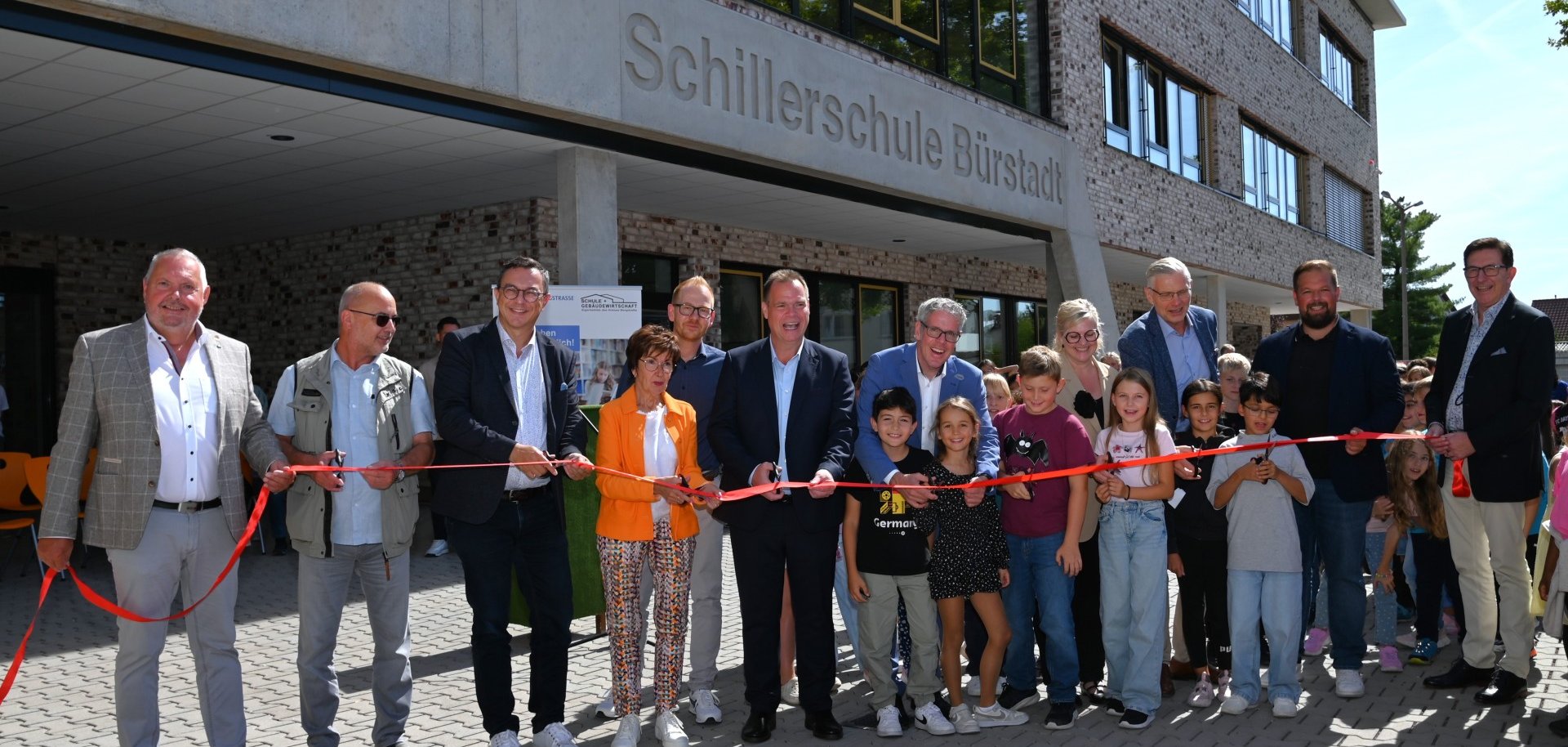 Landrat Christian Engelhardt schneidet gemeinsam mit Vertreterinnen und Vertretern des Schulträgers, Schulamtes, der Schulgemeinde, Baubeteiligten und Kindern ein rotes Band vor dem Schulgebäude durch.