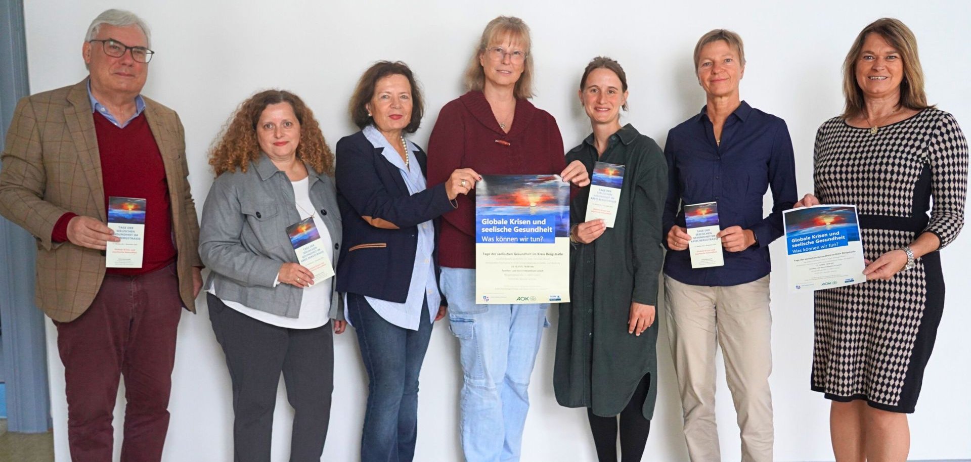 Die Erste Kreisbeigeordnete und Gesundheitsdezernentin Angelika Beckenbach steht gemeinsam mit Jutta Gemeinhardt (Klinikseelsorgerin des Evangelischen Dekanats Bergstraße), Luisa Götzinger (Leiterin des Gemeindepsychiatrischen Zentrums Bergstraße-Ried), Hendrika Mühlbauer (vom Sozialpsychiatrischen Dienst des Kreises Bergstraße), Angela Abel (Geschäftsführerin des Psychosozialen Hilfsvereins Heppenheim e.V.), Fides Matejka (Bereichsleiterin der Regionalen Diakonie Rimbach) sowie Constantin von Gatterburg (Vorsitzender des Helseekon-Klubs Heppenheim e. V) im Flur und halten das Programm der Tage der seelischen Gesundheit in den Händen.