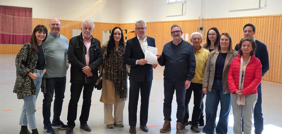 Bescheidübergabe Hallenboden Ober-Mumbach Landrat Christian Engelhardt (5. v. li.) überreichte den Förderbescheid für die Turnhalle in Ober-Mumbach an Mörlenbachs Bürgermeister Erik Kadesch. Mit dabei (v.l.n.r.): Rekha Krings, Geschäftsführerin der Interessengemeinschaft Odenwald (IGO), Gerhard Ohlig, Vorsitzender der Gemeindevertretung Mörlenbach, Andreas Lange, Vorsitzender der SKG Ober-Mumbach, die Kreistagsabgeordnete Anna Maria Schranz, Klaus Zopf, Ortsvorsteher von Ober-Mumbach, Ines Günther-Laake, Regionalmanagerin (LEADER) bei der IGO, Christina Müller, Fachbereichsleiterin Dorf- und Regionalentwicklung in der Kreisverwaltung, Simon Menden, Leiter der Abteilung Ländlicher Raum in der Kreisverwaltung sowie Dorothea Bartossek, Assistenz der Geschäftsleitung bei der IGO.