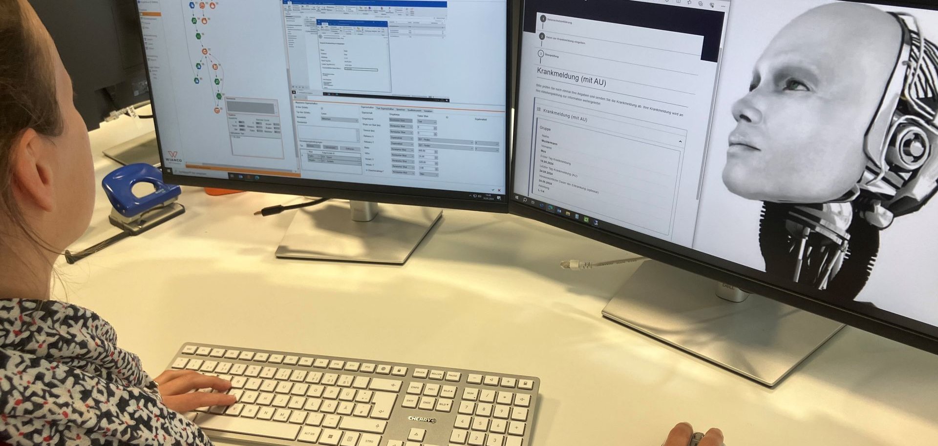 Eine Frau sitzt am Rechner vor zwei Bildschirmen, auf einem ist ein Roboterkopf zu sehen.