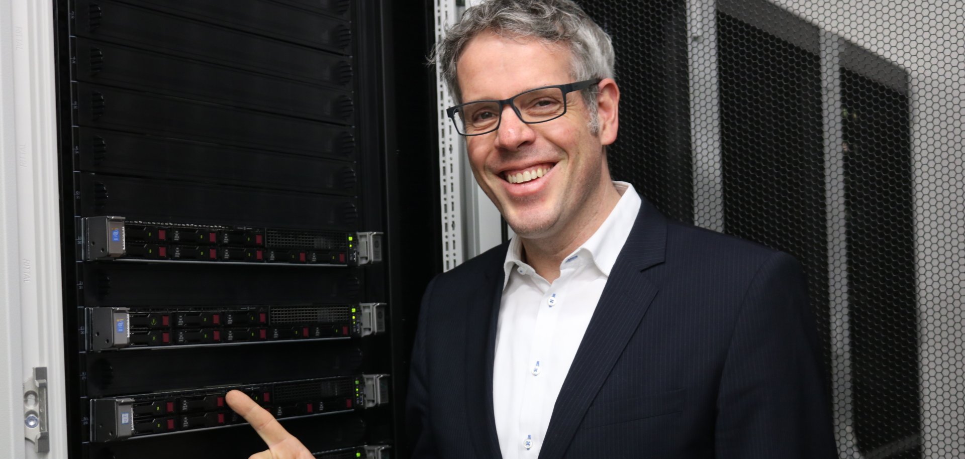 Landrat Christian Engelhardt steht vor einem Server und zeigt lächelnd drauf.