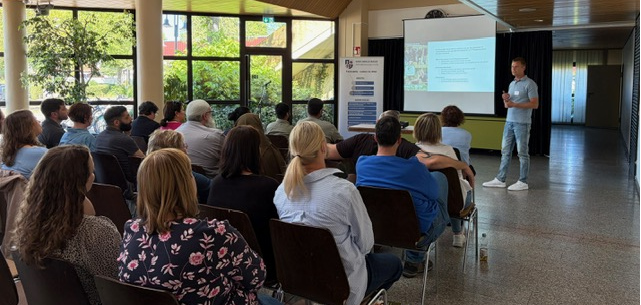 Pflegetisch Ein Mensch trägt eine Powerpoint-Präsentation vor während weitere Mitglieder in mehreren Reihen sitzend zuhören.