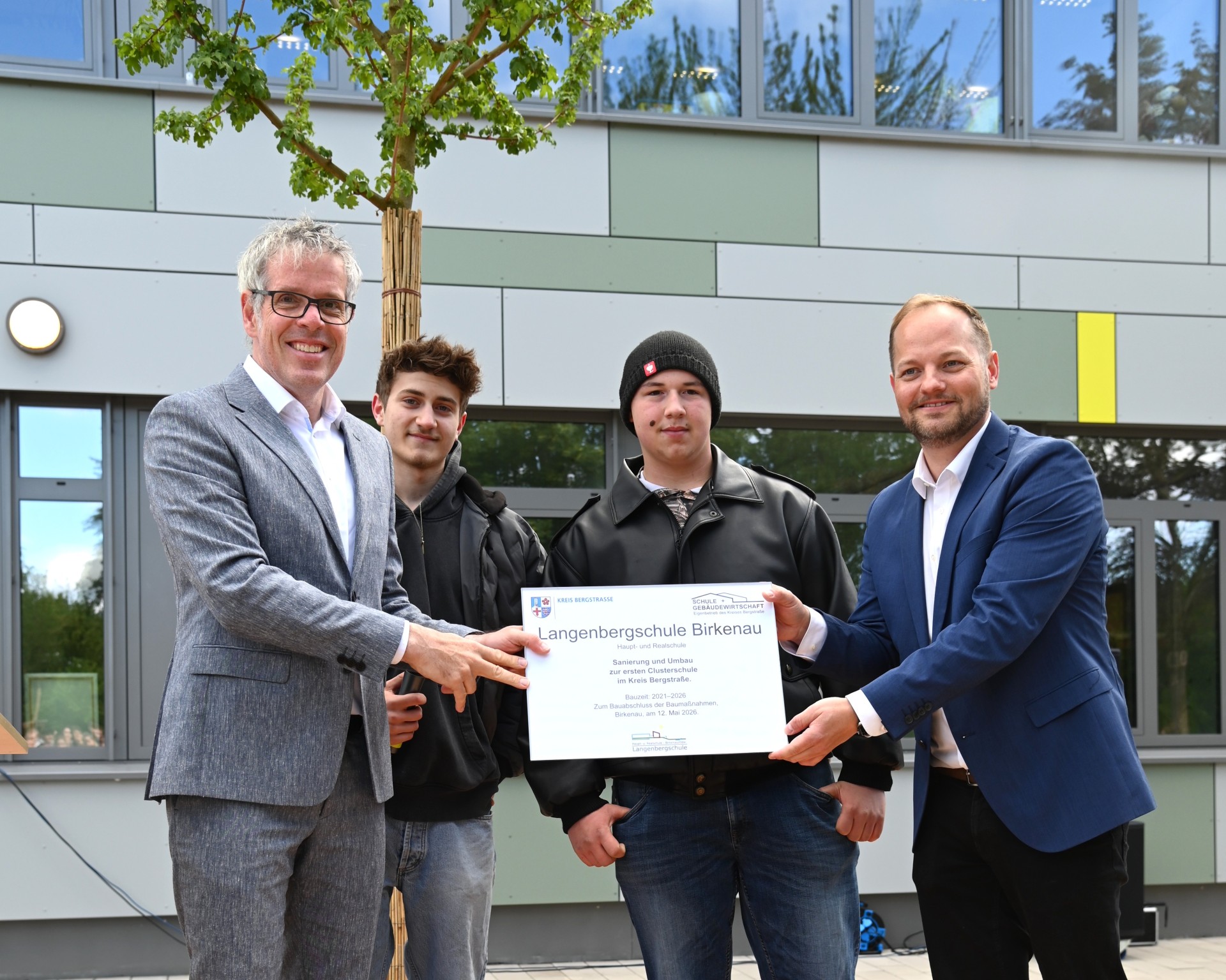 Landrat Christian Engelhardt überreicht dem stellvertretenden Schulleiter, Thomas Rech, sowie den beiden Schulsprechern Hennes und Manuel ein Schild, das an die abgeschlossene Sanierung erinnert.