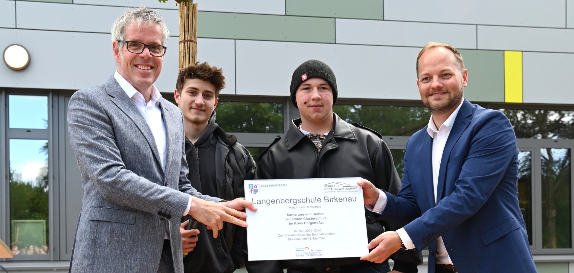 Landrat Christian Engelhardt überreicht dem stellvertretenden Schulleiter, Thomas Rech, sowie den beiden Schulsprechern Hennes und Manuel ein Schild, das an die abgeschlossene Sanierung erinnert.