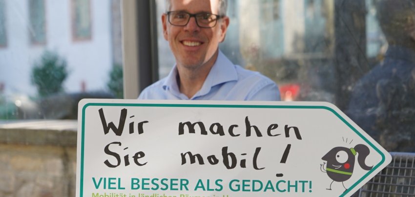 Landrat Christian Engelhardt sitzt in einer Bushaltestelle und hält das Aktionsschild in den Händen.