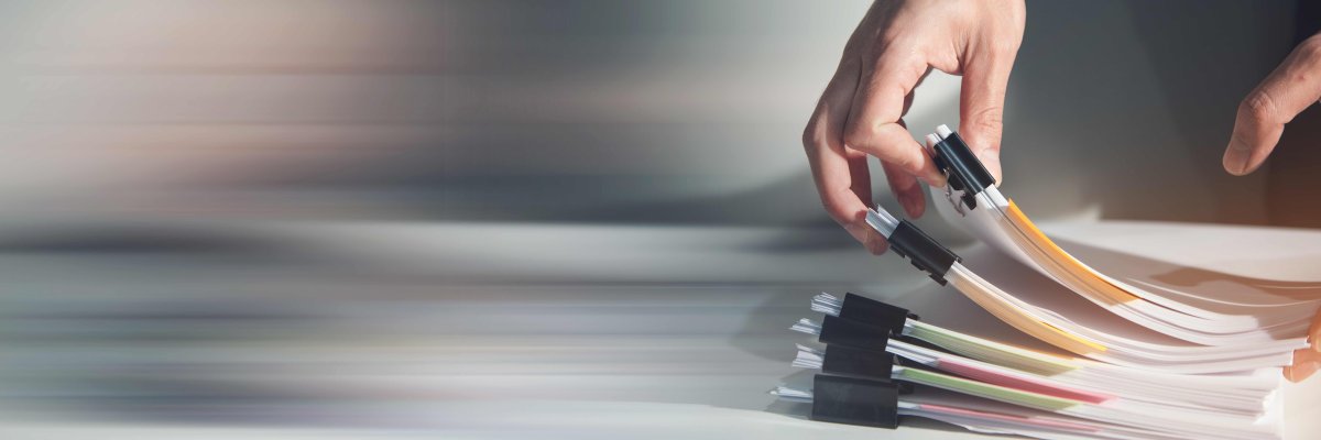 Eine Hand blättert durch zusammengeheftete Papierunterlagen, die auf einem Tisch liegen.