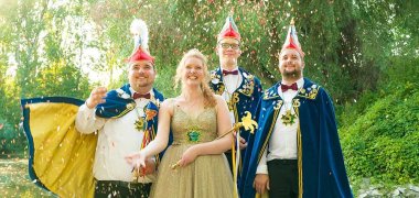 Zu sehen sind drei Männer in dunkelblauen Umhängen und Elferratshüten, in der Mitte steht eine blonde Frau in einem goldenen Kleid mit Krone und Zepter