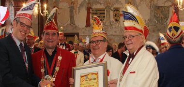 Blick auf einen vollen Saal. Im Vordergrund steht Landrat Christian Engelhardt mit drei weiteren Männern, alle mit Elferratsmützen auf dem Kopf. Es wird ein Fastnachtsorden uns eine gerahmte Urkunde ins Bild gehalten.