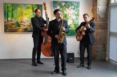 3 Musiker mit Streichinstrumenten spielen vor Gemälden, auf denen die regionale Landschaft abgebildet ist.