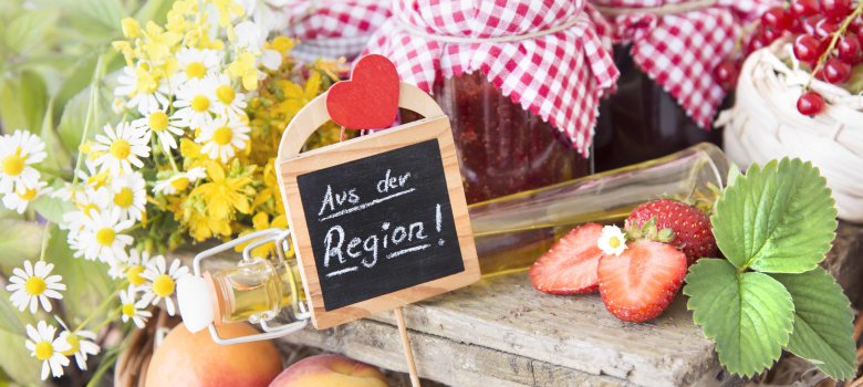 verschiedene regionale Produkte wie Erdbeeren, Äpfel und Marmelade sind zwischen Blumen drapiert. in der Mitte ist ein kleines Holzschild mit der Aufschrift "Aus der Region" zu sehen.