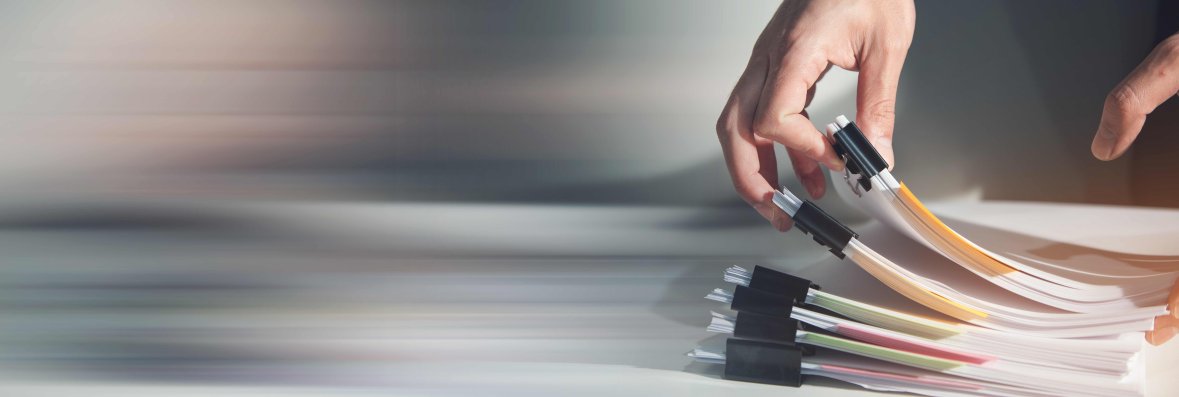 Stapel mit Unterlagen Eine Hand blättert durch zusammengeheftete Papierunterlagen, die auf einem Tisch liegen.