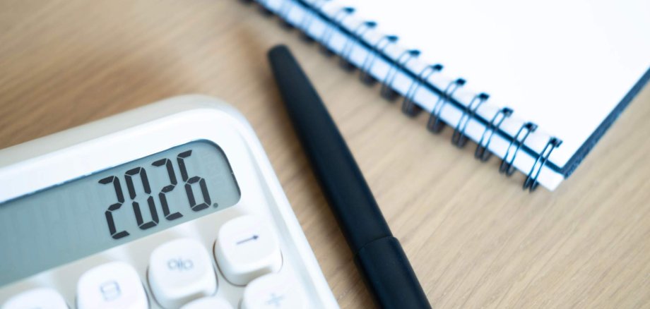 Ein Taschenrechner, auf dem die Zahl 2026 zu sehen ist, liegt neben einem Notizbuch und einem Kugelschreiber.