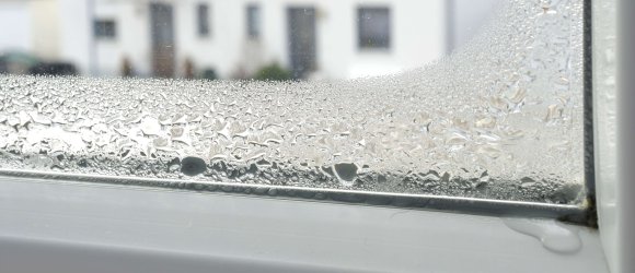An der unteren linken Ecke einer Glasscheibe in einem weißen Fensterrahmen sammelt sich Kondenswasser in großen Tropfen.