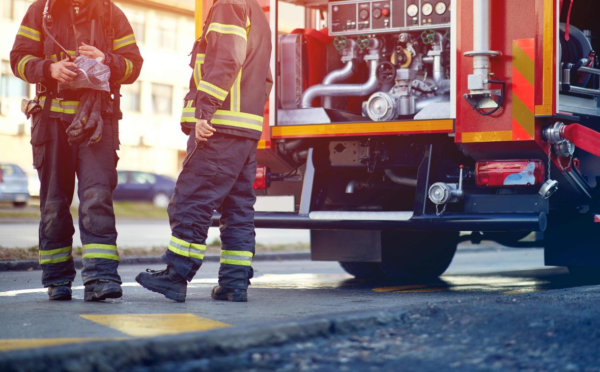 Feuerwehr im Einsatz Man sieht in der rechten Bildhälft die untere Hälfte des Hecks eines professionellen Feuerwehrautos mit der entsprechenden Ausstattung. Links daneben stehen zwei Feuerwehrleute in Schutzkleidung. Diese beiden Personen sind nur von der Schulter an abwärts zu sehen.