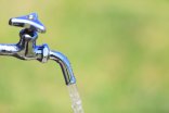 Trinkwasserhahn Das Bild zeigt einen geöffneten Trinkwasserhahn, aus dem frisches Wasser vor einem unscharfen grünen Hintergrund fließt.