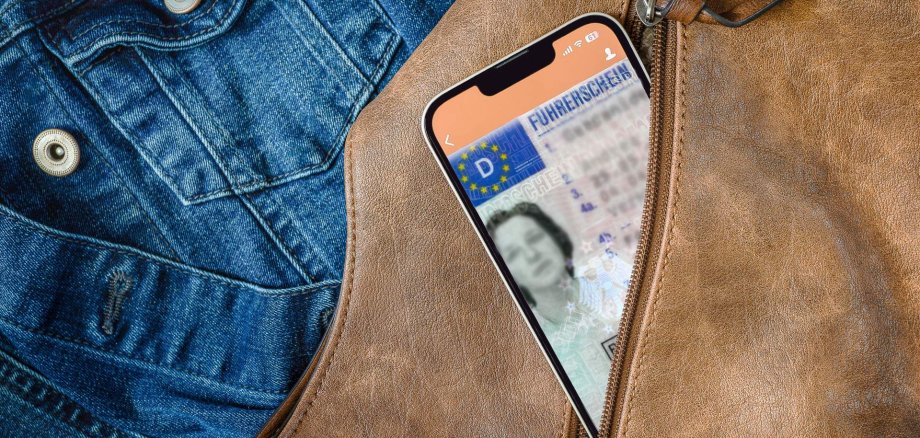 Digitale Führerschein-Dienste Aus einem geöffneten Reißverschluss einer braunen Handtasche schaut ein Smartphone heraus, auf dem gerade eine App mit digitalem Führerschein geöffnet ist. Im Hintergrund ist eine Jeansjacke zu sehen,