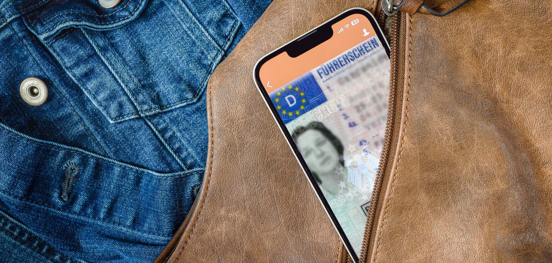 Aus einem geöffneten Reißverschluss einer braunen Handtasche schaut ein Smartphone heraus, auf dem gerade eine App mit digitalem Führerschein geöffnet ist. Im Hintergrund ist eine Jeansjacke zu sehen,