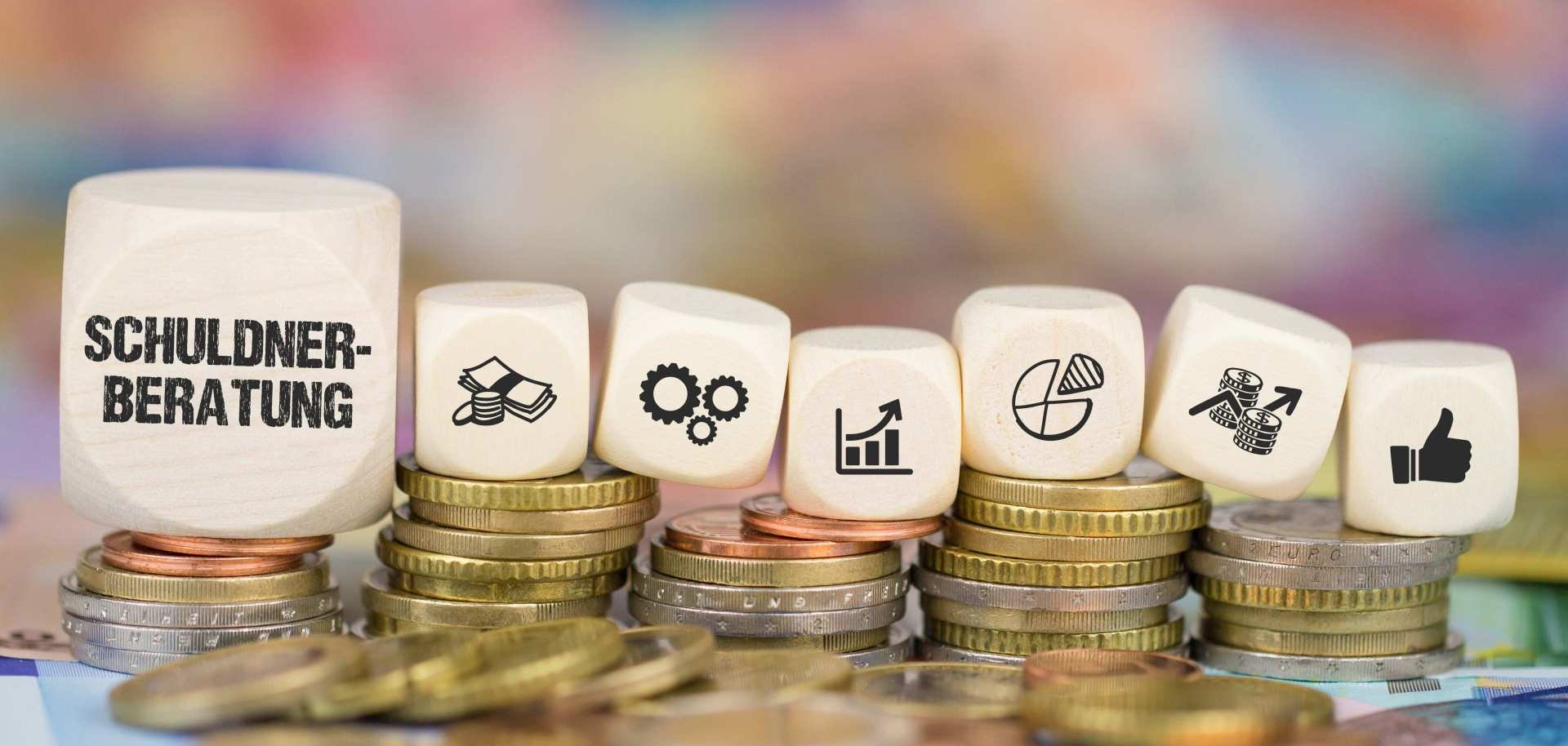 Ein paar Würfel mit verschiedenen Symbolen und dem Wort "Schuldnerberatung" liegen auf Stapeln aus Münzen.