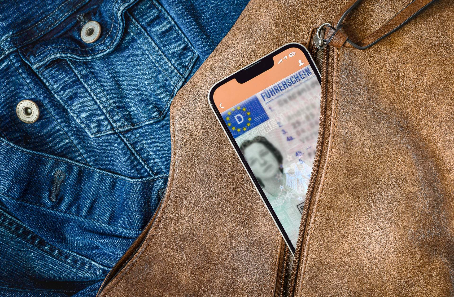 Aus einem geöffneten Reißverschluss einer braunen Handtasche schaut ein Smartphone heraus, auf dem gerade eine App mit digitalem Führerschein geöffnet ist. Im Hintergrund ist eine Jeansjacke zu sehen,