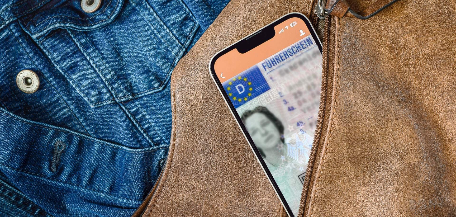 Aus einem geöffneten Reißverschluss einer braunen Handtasche schaut ein Smartphone heraus, auf dem gerade eine App mit digitalem Führerschein geöffnet ist. Im Hintergrund ist eine Jeansjacke zu sehen,