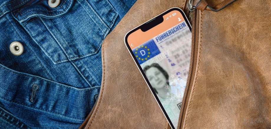 Onlinedienste Führerschein Aus einem geöffneten Reißverschluss einer braunen Handtasche schaut ein Smartphone heraus, auf dem gerade eine App mit digitalem Führerschein geöffnet ist. Im Hintergrund ist eine Jeansjacke zu sehen,