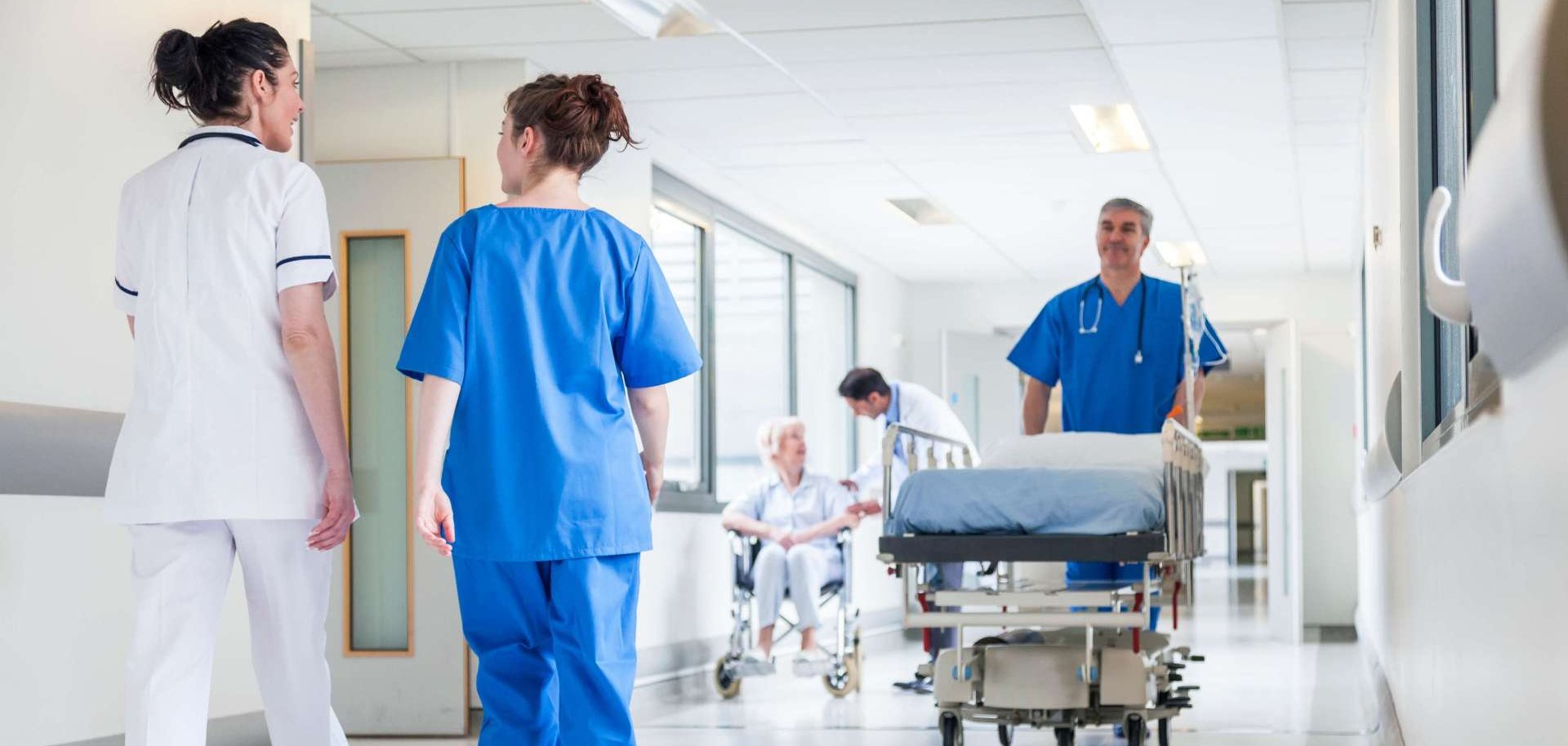 Das Bild zeigt einen Krankenhausflur: Ein Pfleger schiebt ein Bett, eine Pflegerin unterhält sich mit einer Ärztin, eine Arztkümmert sich um eine ältere Frau im Rollstuhl.
