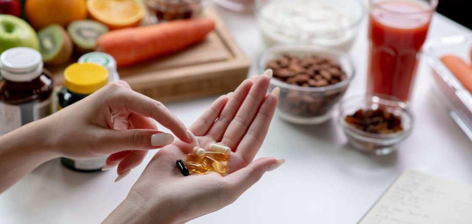 Nahrungsergänzungsmittel Das Bild zeigt die Hand einer Frau, in der sich mehrere Nahrungsergänzungsmittel in Form von Pillen befinden. Der Hintergrund des Bildes zeigt gesunde Lebensmittel wie Gemüse und Obst.