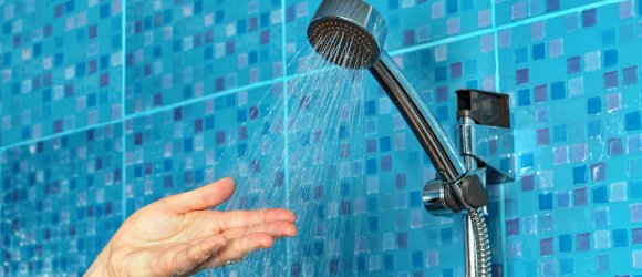 Ein Hand fühlt die Wassertemperatur eines laufenden Duschkopfs vor eine blau gefliesten Wand.