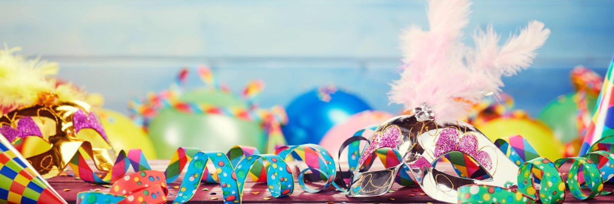 Auf einem Holztisch liegen bunte Luftschlangen und Konfetti. Im Hintergrund sieht man verschwommen bunte Luftballons.