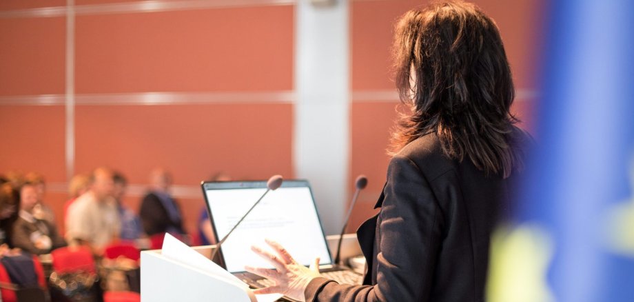 Eine Politikerin steht in einem Saal und spricht vor einem Mikrofon und spricht zu den Anwesenden.